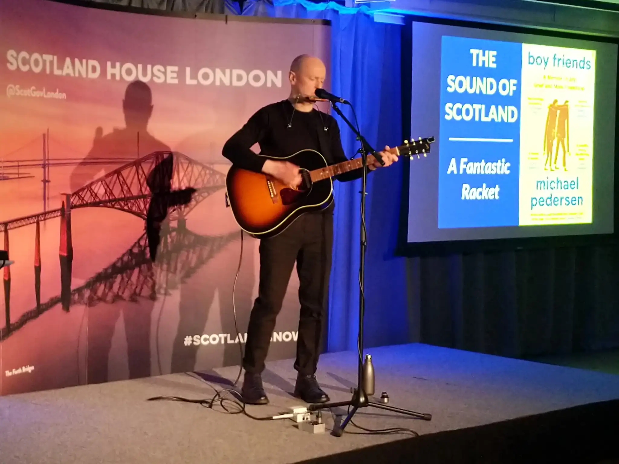 A man, dressed all in black, plays an acoustic guitar on stage, and leans
forward to sing into the microphone. Behind him, banners read 'Scotland House
London' and 'The Sound of Scotland: A Fantastic Racket'.