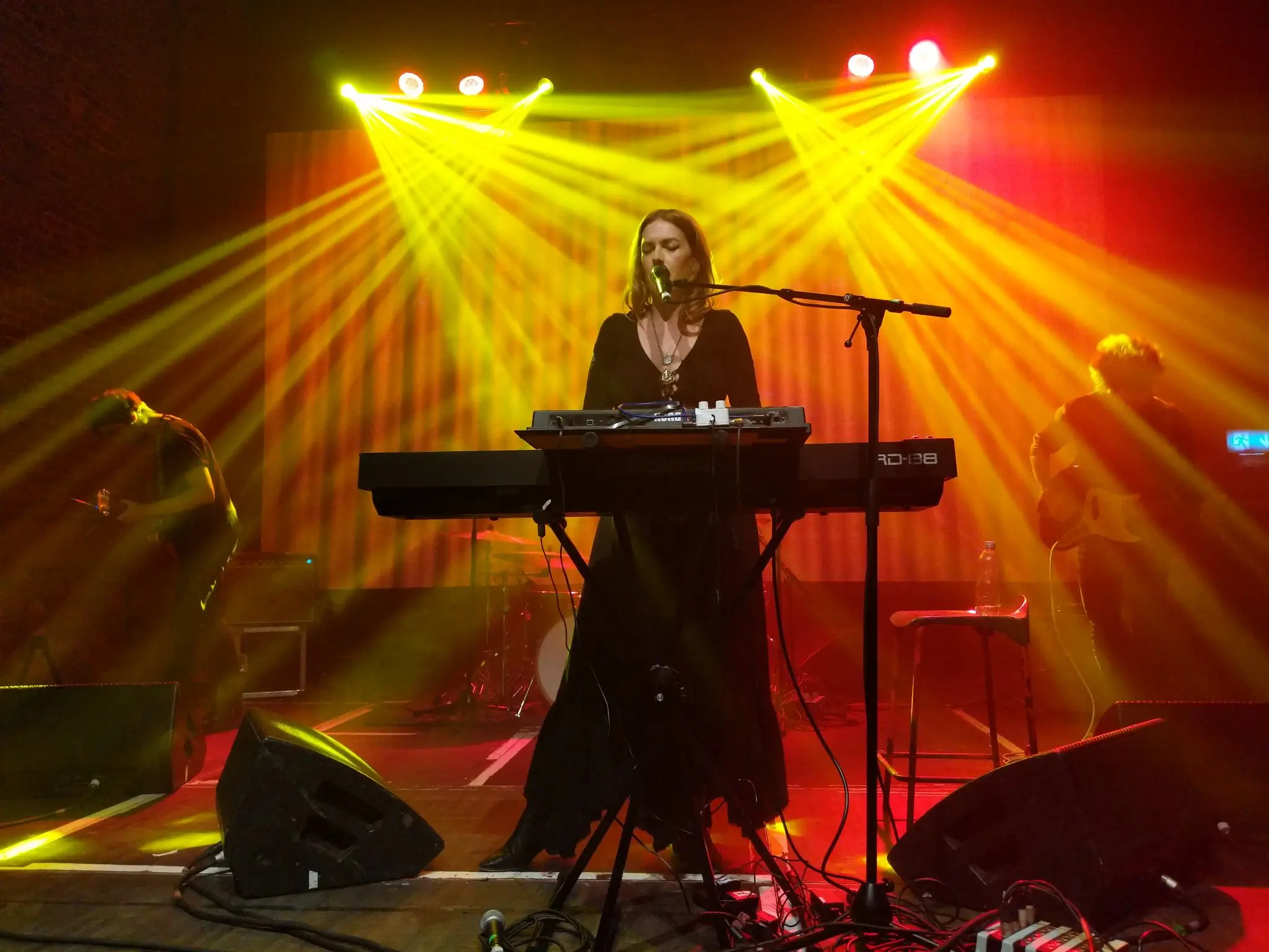 A woman is standing in the middle of the stage behind a couple of keyboards, with orange and yellow lights radiating behind her. A guitarist and bassist are visible on each side, further back, moving energetically.