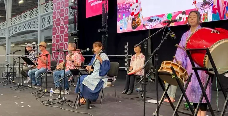 A row of performers on a stage variously playing the sanshin, the drum,
and singing