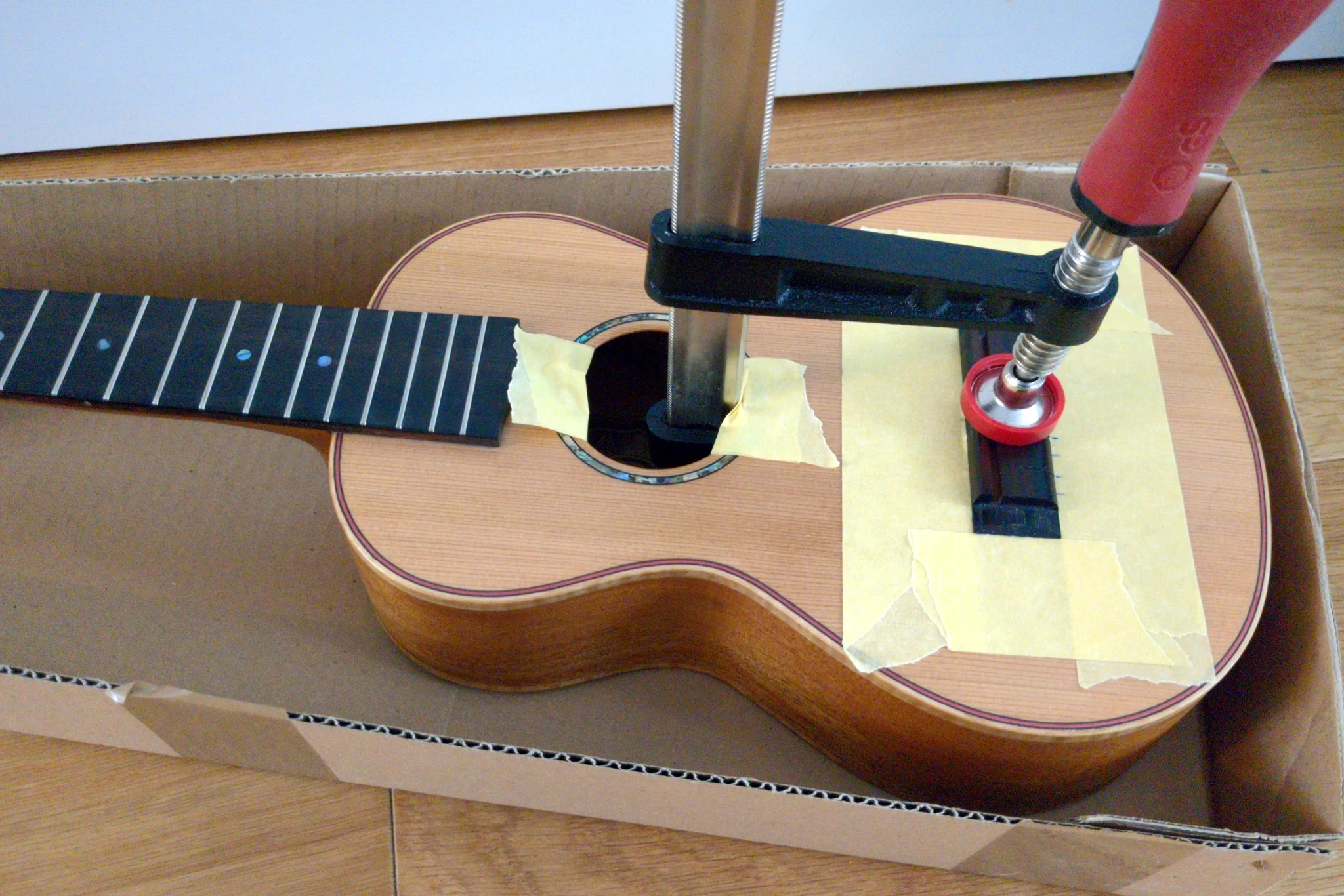 The same ukulele. It's sitting in a cardboard box on the floor. There
is masking tape around the sound hole for protection, around the bridge to
locate it, and a bit G clamp is holding the bridge in place.