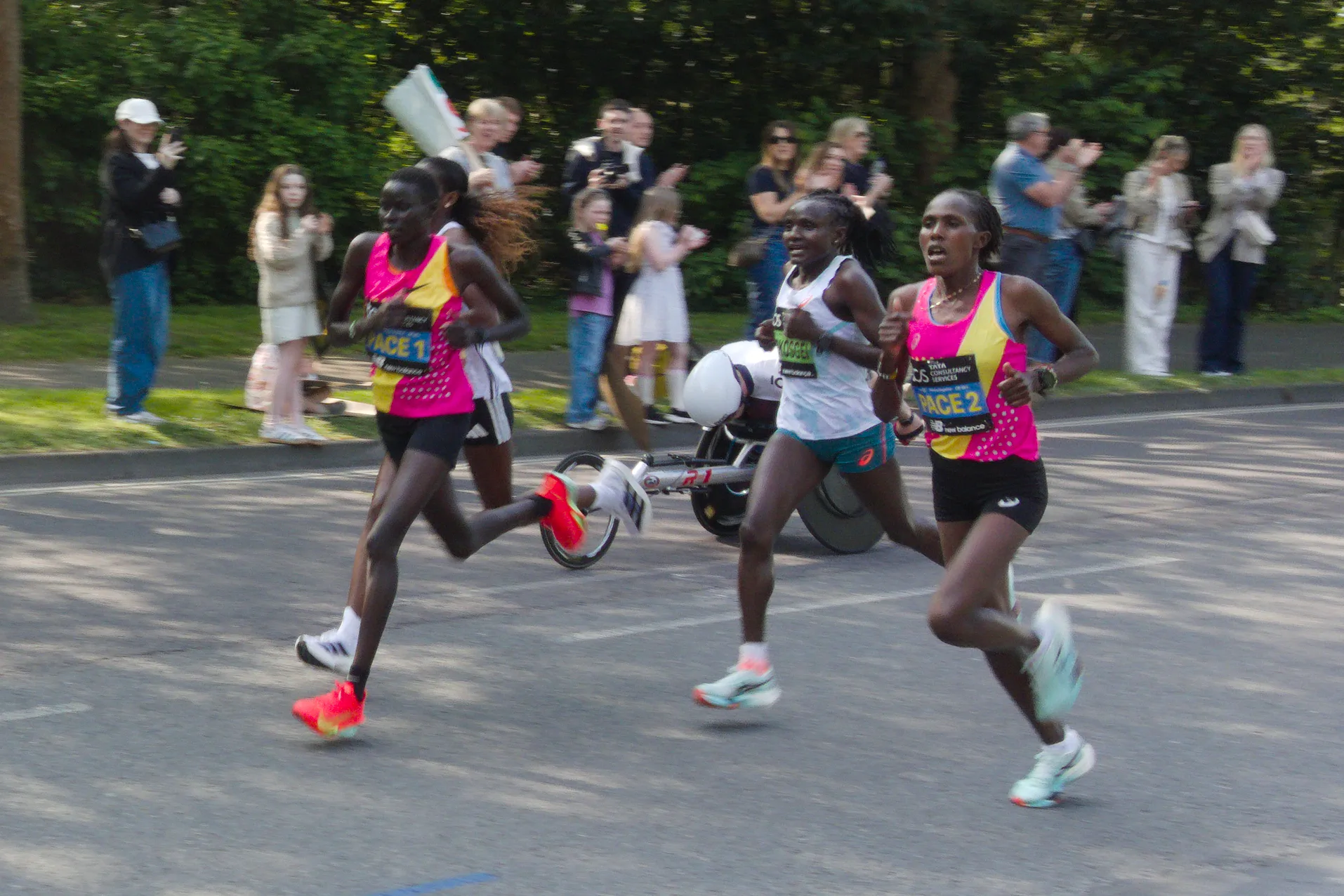 Two women running a marathon together with their pacers. A wheelchair
racer passes by in the background.