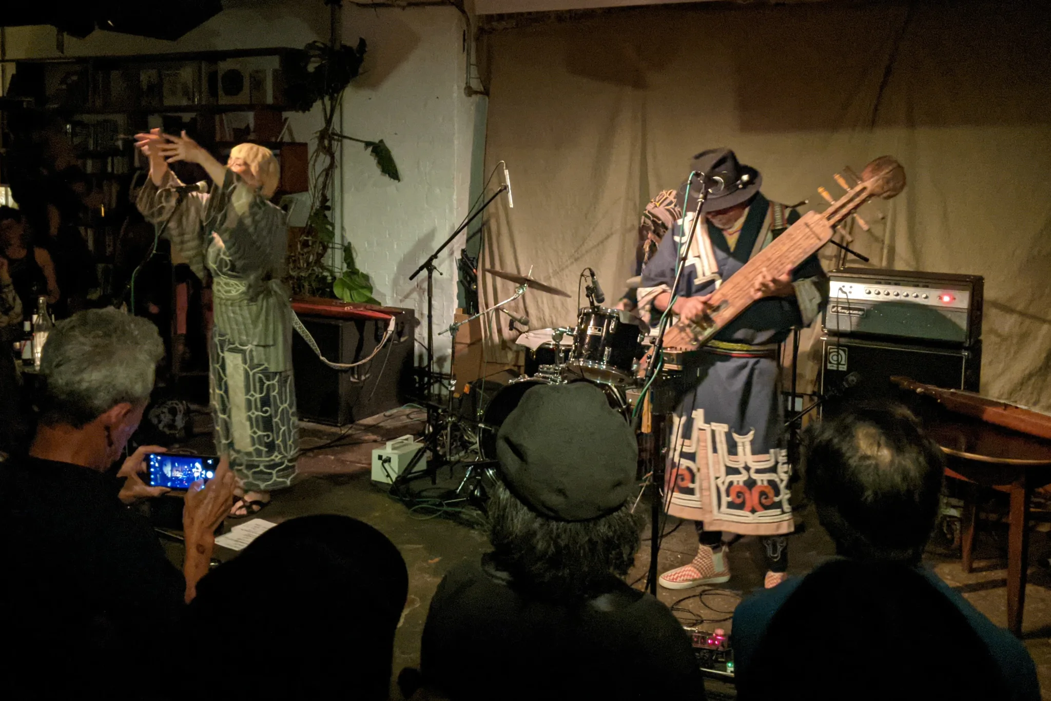 Musicians in Ainu dress on stage. There is a drummer at the back. On the left, a singer has her arms raised. On the right, a man plays the tonkori.