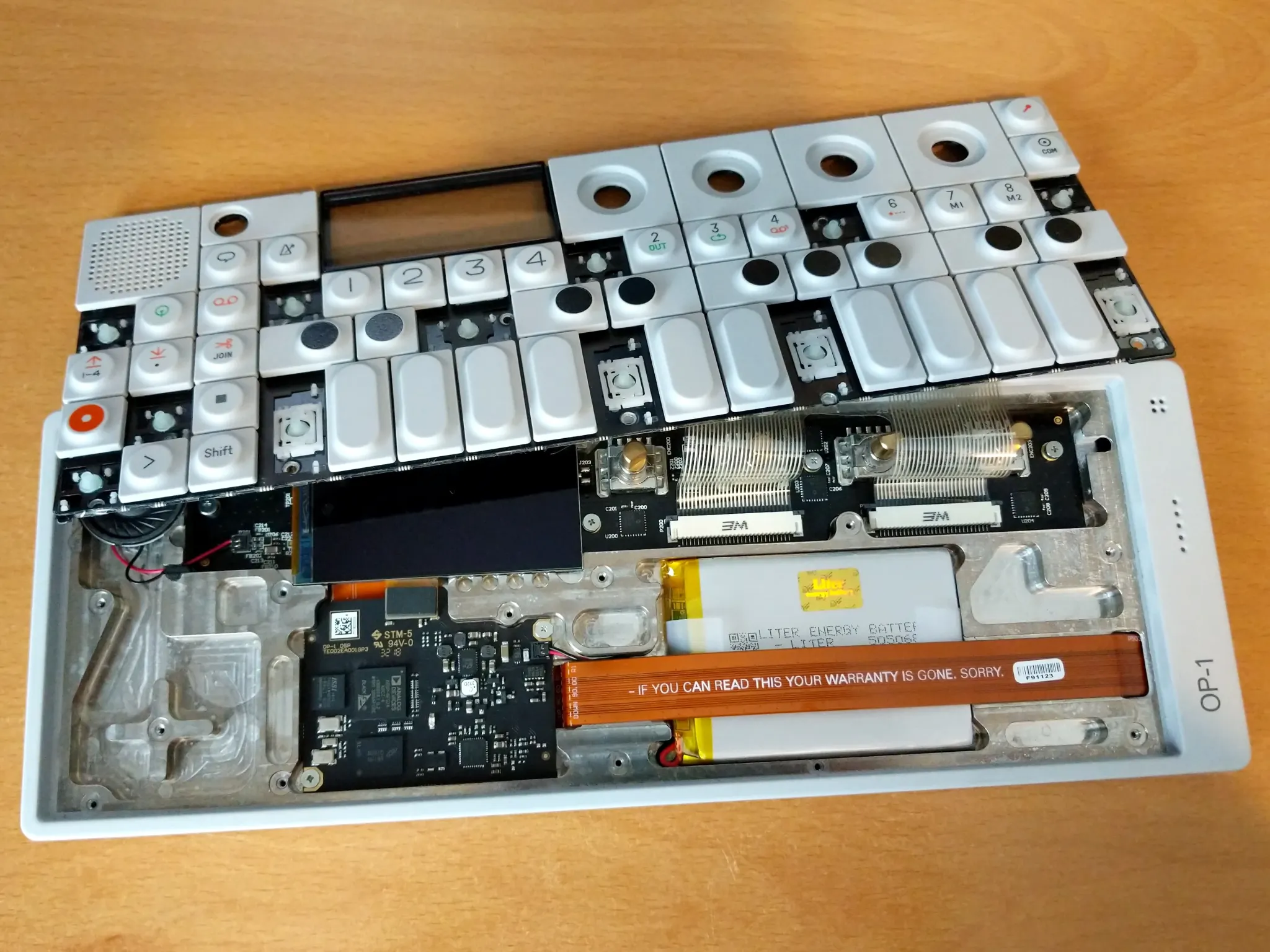 A partially disassembled OP-1. The keyboard is moved up to expose the
internal circuitry, and a cable on which is written, 'IF YOU CAN READ THIS YOUR
WARRANTY IS GONE. SORRY'