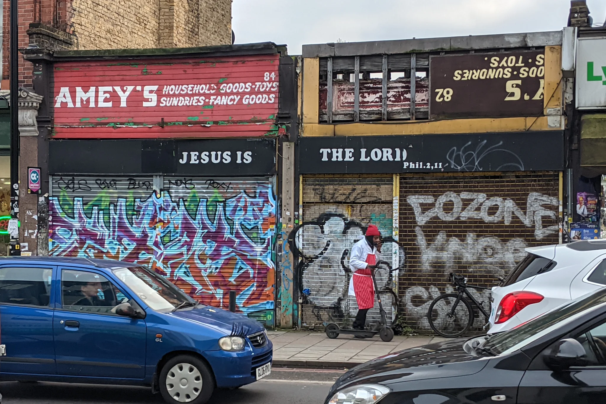 On the far side of the road, beyond passing cars, is a shuttered shop.
It's covered with graffiti, and the remnants of an old sign are visible.
Directly above the frontage is a newer sign, reading JESUS IS THE LORD.
Someone in a red hat and apron is trundling along the pavement on a scooter.