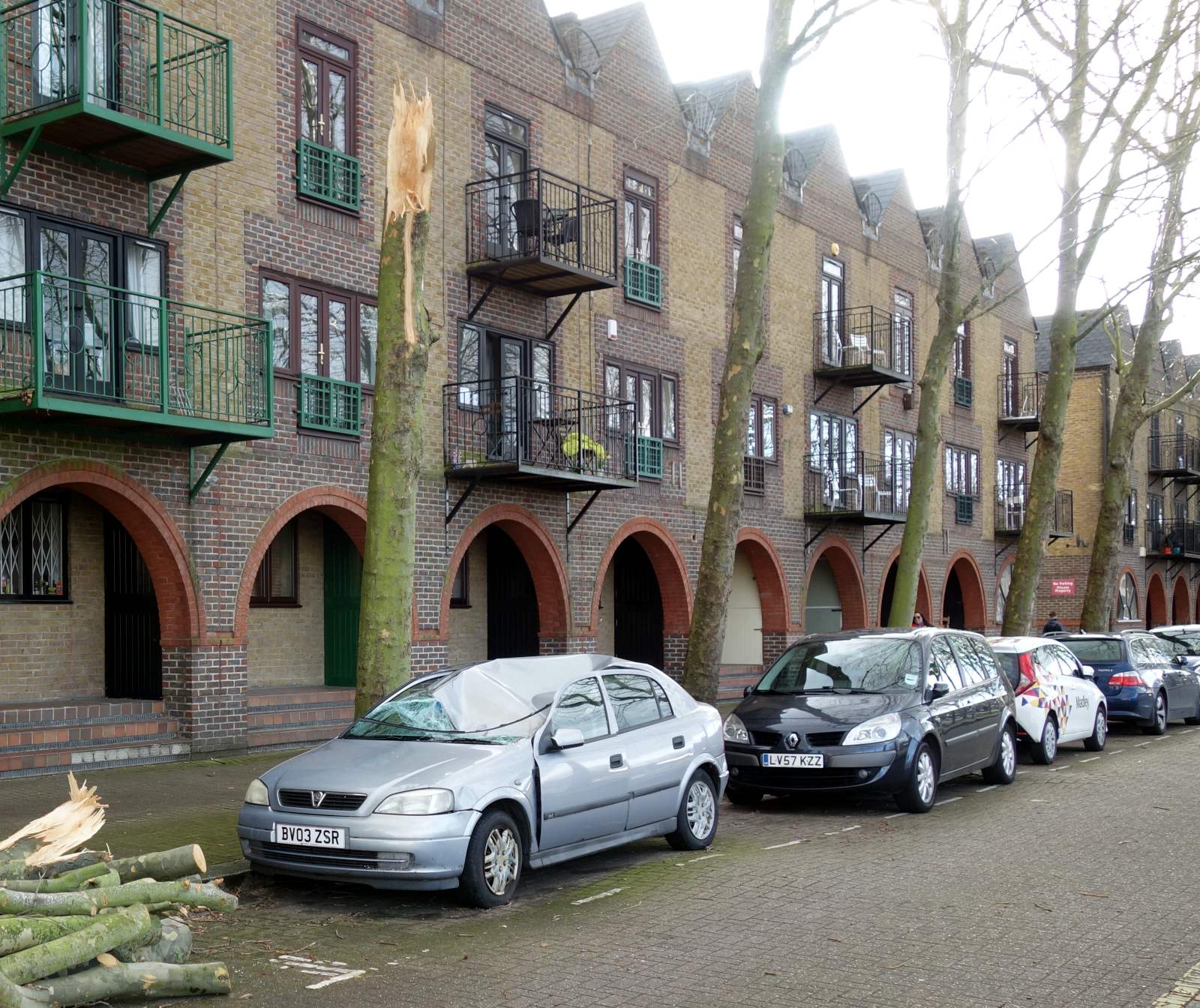 A car is parked on the side of the road next to a tall tree and a row of
buildings. The tree is snapped off at about third floor height, and the
roof of the car is crushed down to below the bonnet height.