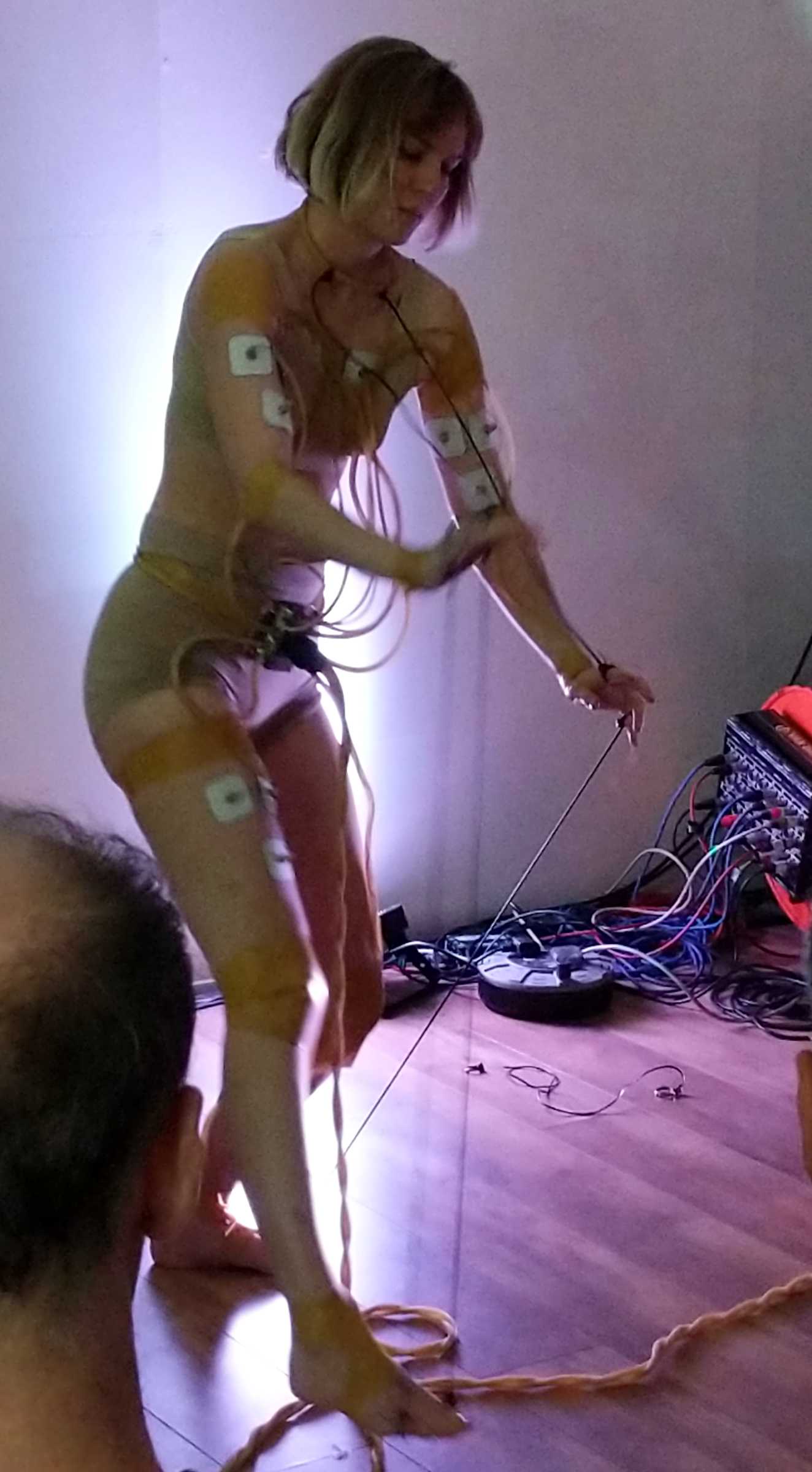 A woman with muscle sensors attached to her arms and legs, stretch sensors
from fingers to toes, and cables everywhere. She is moving as she uses the
sensors to control a musical performance.