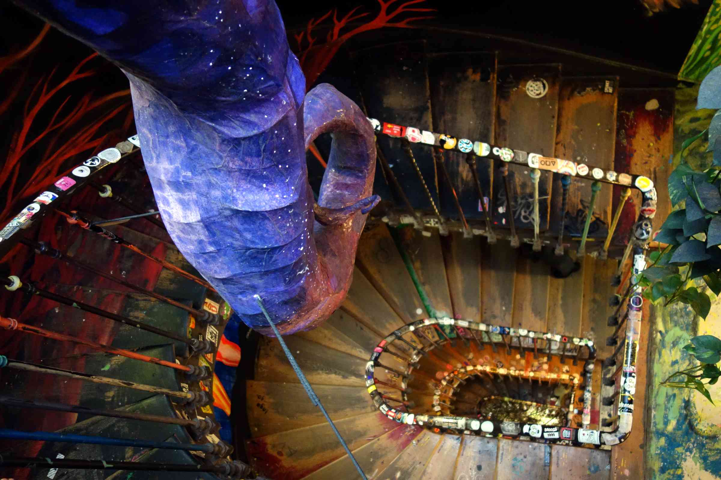 A giant purple tentacle hangs in the centre of a stairwell. The stairs curve round and round below. The stairs and walls are covered in painted designs and stickers.